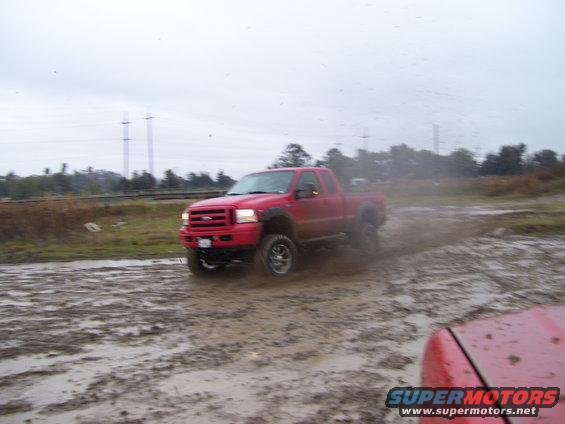 Red pickup truck driving through mud; another red vehicle is partially visible in the foreground