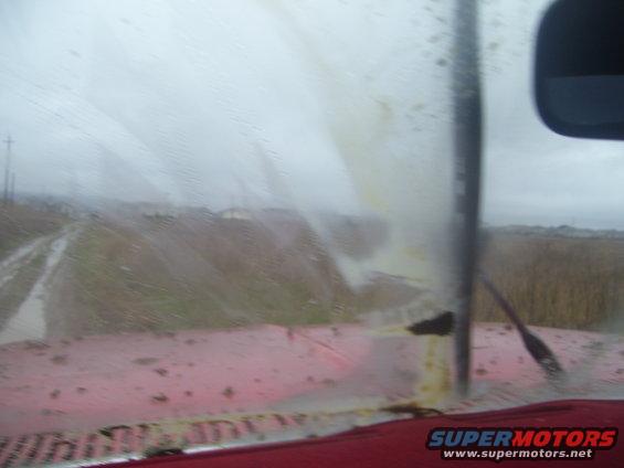 view from inside a vehicle looking out a dirty windshield at a muddy road and fields