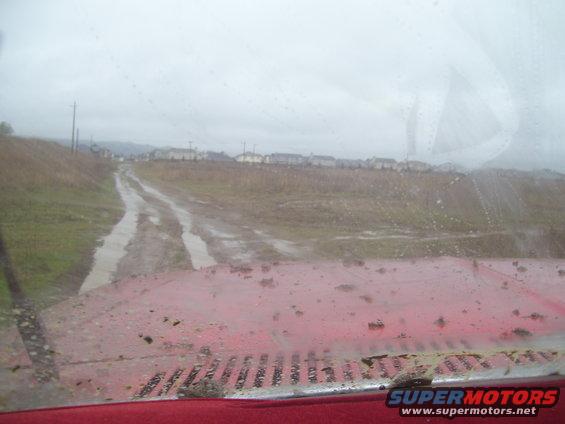 Red vehicle hood covered in mud, viewed from inside through a dirty windshield looking at a muddy road