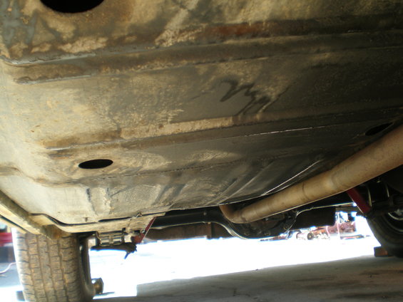 Underside of a vehicle showing the exhaust pipe and rear axle. The frame appears to have some rust