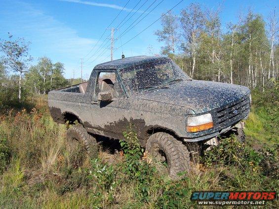 Mud-covered dark green Ford Bronco, 3/4 front view, stuck in tall grass and mud