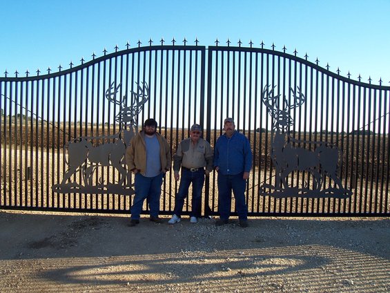 Three men stand before a tall, ornate black metal gate large deer silhouettes on either side