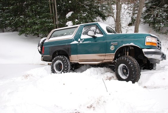 Green 1996 Ford Bronco with tan trim driving through deep snow in a wooded area