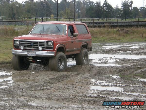 1986 Ford Bronco driving through a muddy field. License plate reads 560E398