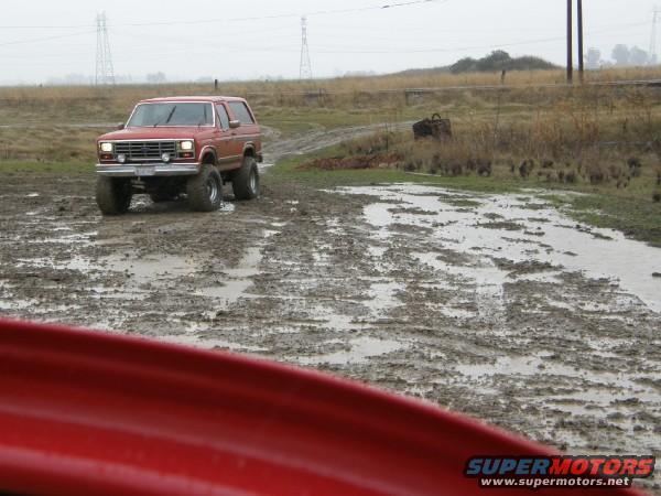 Red Ford Bronco with lifted suspension driving on a muddy track. The Bronco has a removable top