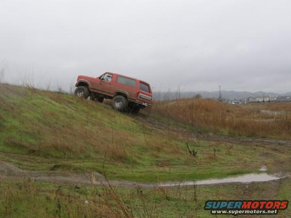 Red 1987 Ford Bronco climbing a muddy, grassy hill with a cloudy sky