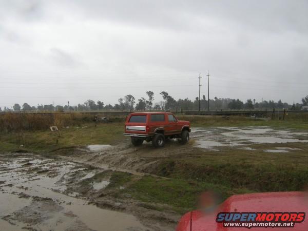 Red SUV driving through muddy field. Another red vehicle is in the foreground