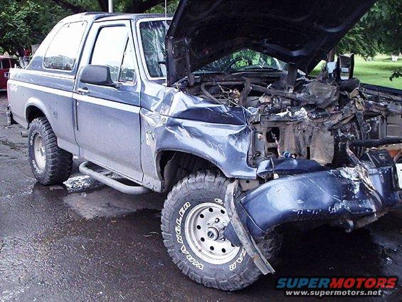 Front-end damaged Ford Bronco with hood open. The front bumper is detached and the grille is gone