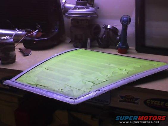 Car window pane covered in green masking tape, resting on a workbench with tools and parts in the background