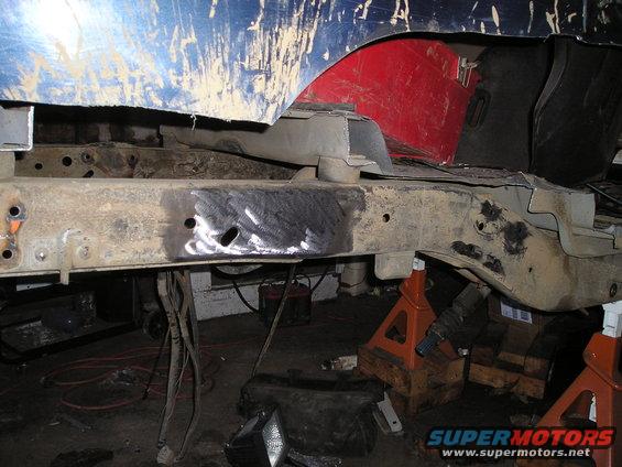 Vehicle frame with rust and repair patches, supported by jack stands. Blue and red body panels are visible above
