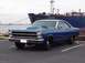 1966 Ford Fairlane - A blue coupe with a prominent hood scoop is parked in front of a large cargo ship.