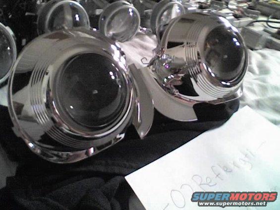 Pair of chrome projector headlights, one partially disassembled. Paper with writing and a SuperMotors logo is in the foreground