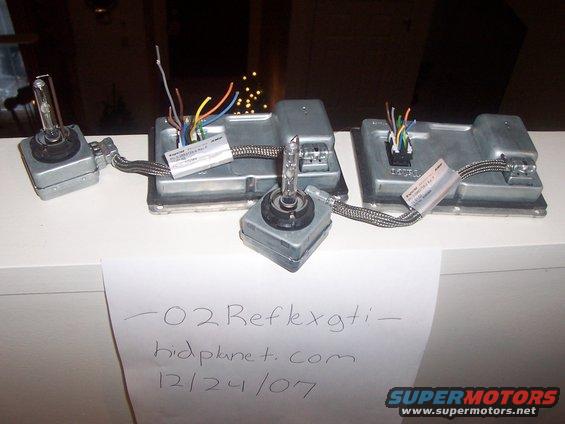 Three HID ballasts and bulbs on a white surface with handwritten notes and a SuperMotors logo