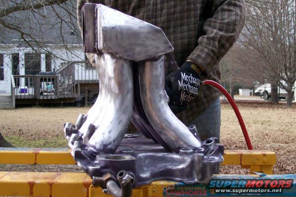 Close-up of a metal intake manifold part with a red hose attached, held by a person wearing gloves. SuperMotors logo visible