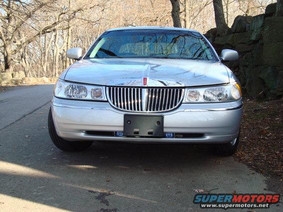 Front view of a silver 2002 Lincoln Town Car parked on a road with trees and a stone wall behind it