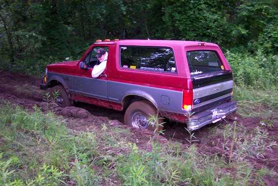 Red and grey Ford Bronco stuck in mud, driver visible, trees and grass in background