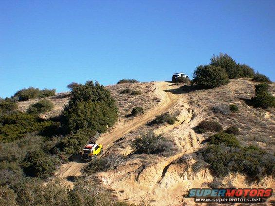Two SUVs, one yellow and one white, driving up a steep dirt trail on a sunny day