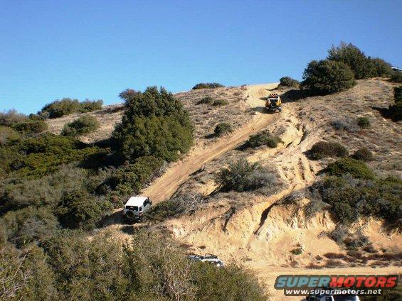 Two Toyota FJ Cruisers and a yellow Jeep ascend a steep, dusty mountain trail under a clear blue sky