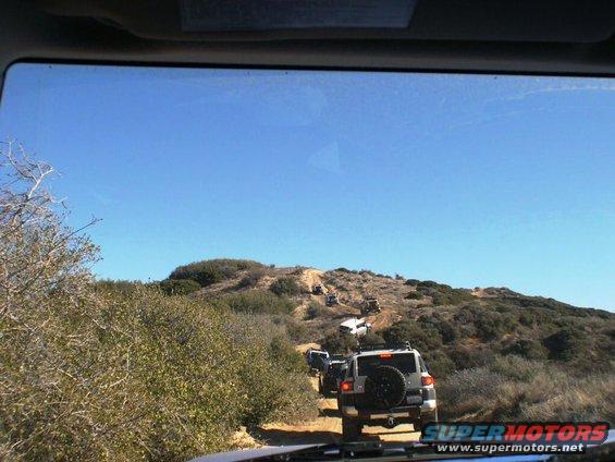 Several Toyota FJ Cruisers driving up a dirt hill off-road. The lead vehicle is silver with a spare tire on the back