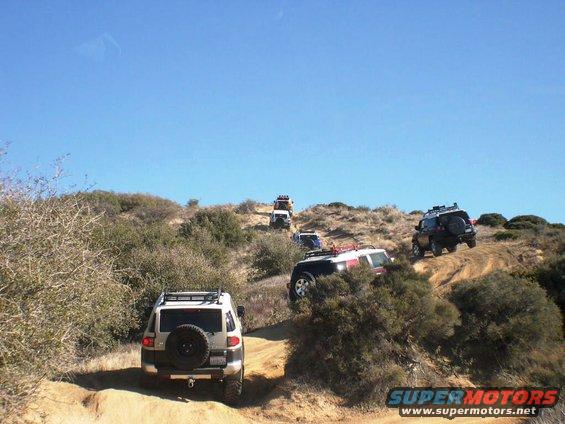 Several Toyota FJ Cruisers driving up a dusty hill on an off-road trail