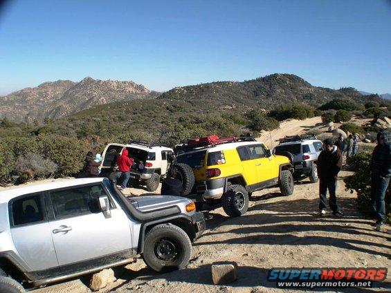 Several Toyota FJ Cruisers parked on a dirt road with mountains in the background. One is yellow, others are white and silver