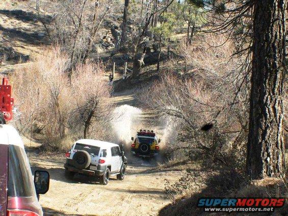 Two 2007 Toyota FJ Cruisers driving through a puddle on a dirt trail, one white and one yellow