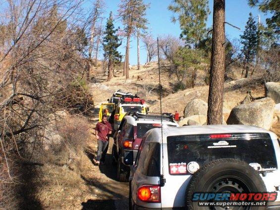 Line of FJ Cruisers driving on a dirt road through a wooded, rocky area. One person stands next to the lead vehicle