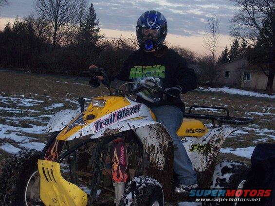Yellow and white ATV with 'Trail Blazer' graphics, covered in mud, with a rider wearing a blue helmet