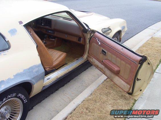 Driver's side interior of a 1979 Chevrolet Camaro, showing tan seats and door panel. Exterior shows primer on the fender