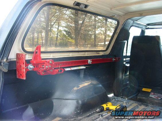 Red hi-lift jack mounted to interior panel of a vehicle. Drill and toolbox visible on the floor