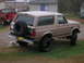 1996 Ford Bronco - A tan 1996 Ford Bronco is parked on grass next to a red pickup truck, with a cat walking near the rear bumper.