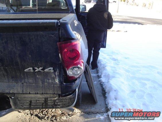 Dark blue pickup truck rear with damaged taillight and loose bumper cover, person standing by open door in snow