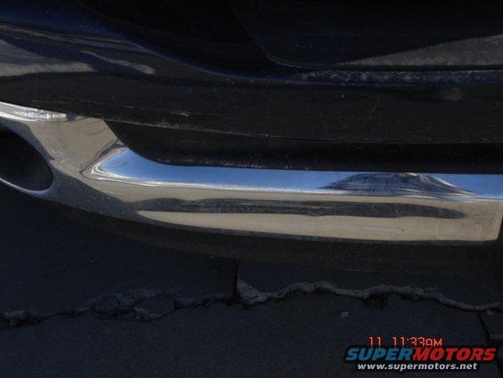 Close-up of a chrome bumper with an exhaust pipe visible on the left. The reflection on the bumper shows a landscape