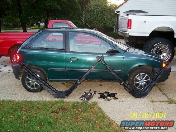 Green hatchback car with leaf springs and hardware on driveway, red truck and white truck in background