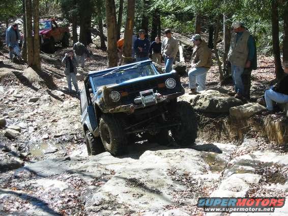 1976 Ford Bronco climbing over rocks on a wooded trail with people watching