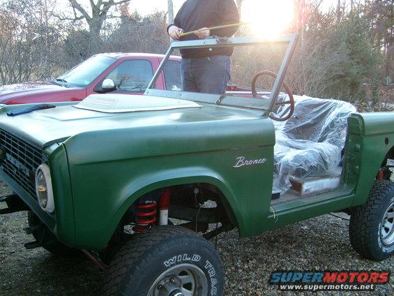 Green Ford Bronco with a person measuring the windshield frame. Red pickup truck in background