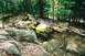 1976 Ford Bronco - A yellow off-road vehicle navigates a challenging rocky path in a wooded area.