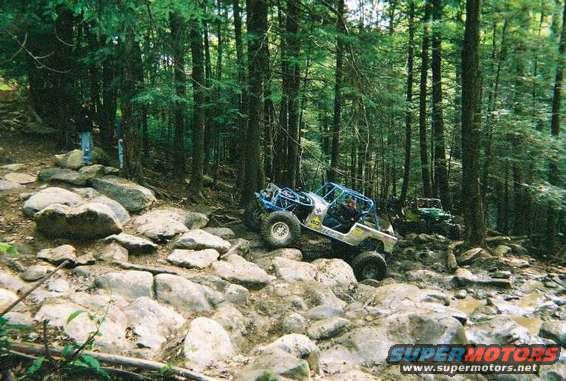 Two off-road vehicles on a rocky, wooded trail. One is in the foreground, the other further back