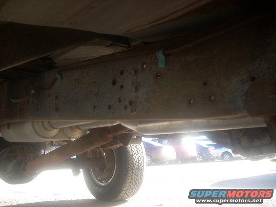 Underside of a rusty vehicle frame with exhaust and rear axle visible. Other vehicles are blurred in the background