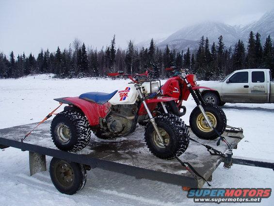 Two Honda SX three-wheelers on a trailer in the snow, with a pickup truck and mountains in the background