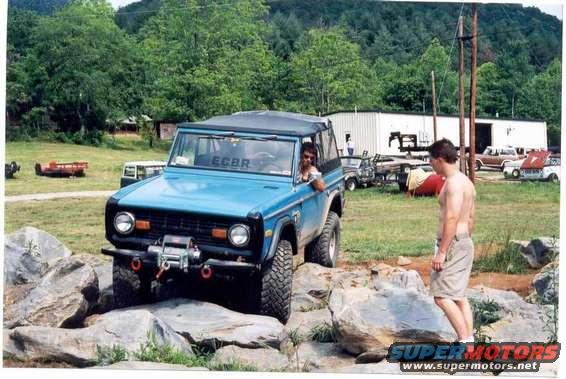 Blue 1976 Ford Bronco navigating rocks on an off-road trail. A man stands nearby
