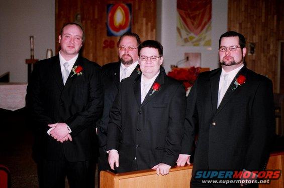 Four men in black suits with red boutonnieres stand in an indoor setting