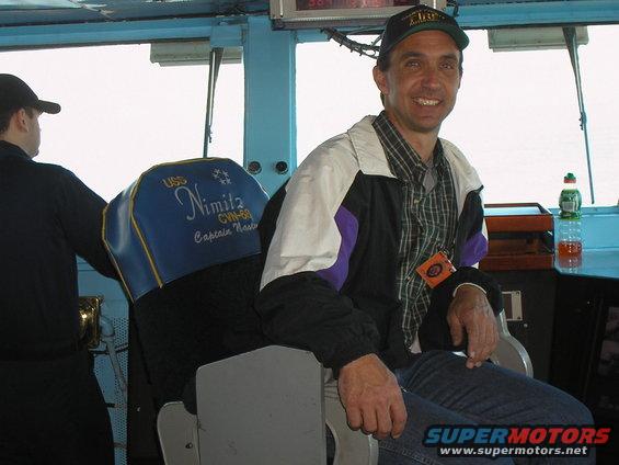 Man in cap and jacket sits in captain's chair on USS Nimitz bridge