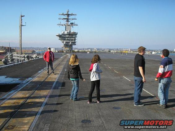 People stand on an aircraft carrier deck with the island structure visible