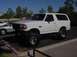 1992 Ford Bronco - A lifted 1992 Ford Bronco is parked in a lot next to a silver sedan under a clear sky.