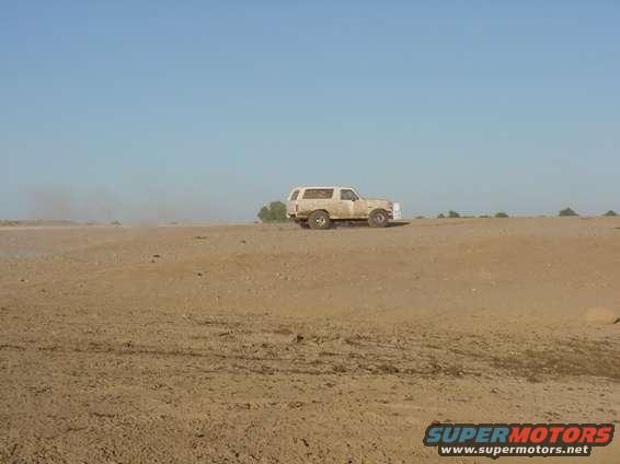 White 1992 Ford Bronco driving on a dirt road in a dry, open area under a clear blue sky