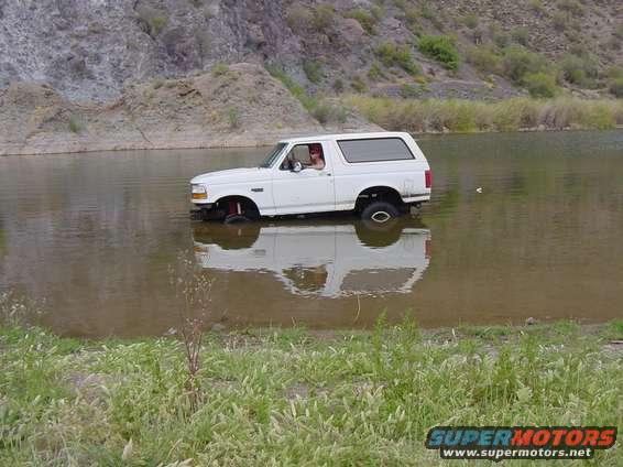 white 1992 Ford Bronco driving through shallow water with a rocky hill in the background