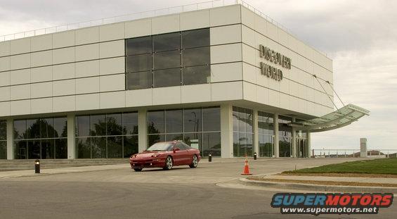 Red 1993 Ford Probe driving in front of a modern white building with "DISCOVER WORLD" signage