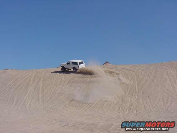 White 1992 Ford Bronco kicking up dust while driving on a sand dune under a clear blue sky