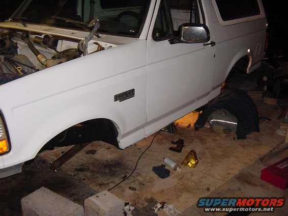 White 1992 Ford Bronco on jack stands, front fender and engine bay visible. Someone is working underneath